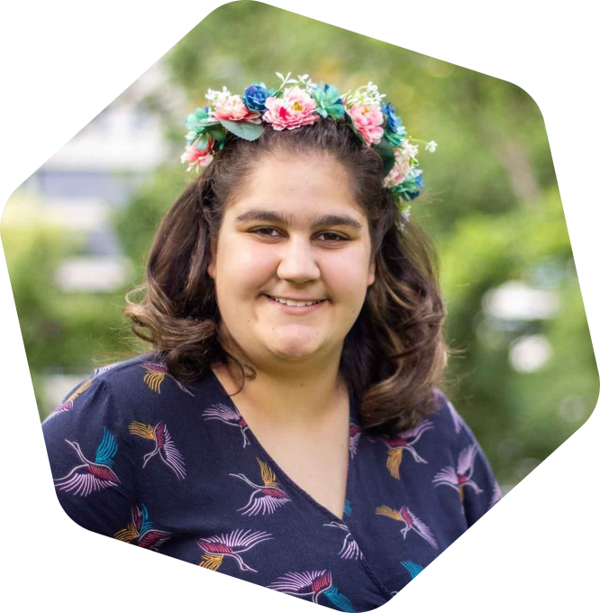 Fel smiling. She is outside amongst greenery and is wearing a flower crown.